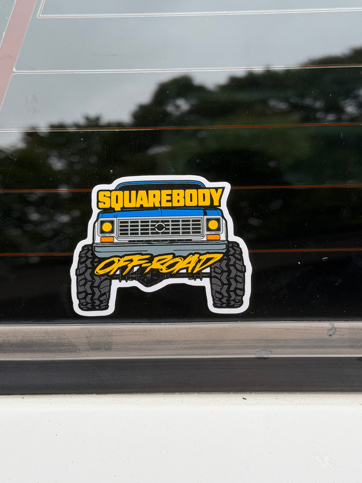 A die-cut vinyl sticker of a blue liftedoff-road vehicle with the words 'SQUAREBODY' and 'OFF-ROAD' in yellow, shown applied to a dark tinted car window.